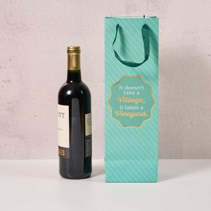 A bottle of red wine sits beside the Drinks On Me Vineyard Wine Bag—a teal reusable bag with handles and gold text, “It doesn’t take a Village, it takes a Vineyard”—a funny hostess gift.