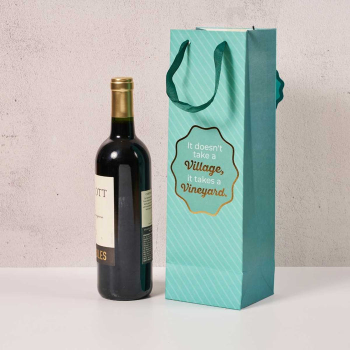 A bottle of red wine stands beside the Drinks On Me Vineyard Wine Bag, featuring green handles and “It doesn’t take a village, it takes a vineyard” on the front—an ideal hostess gift.
