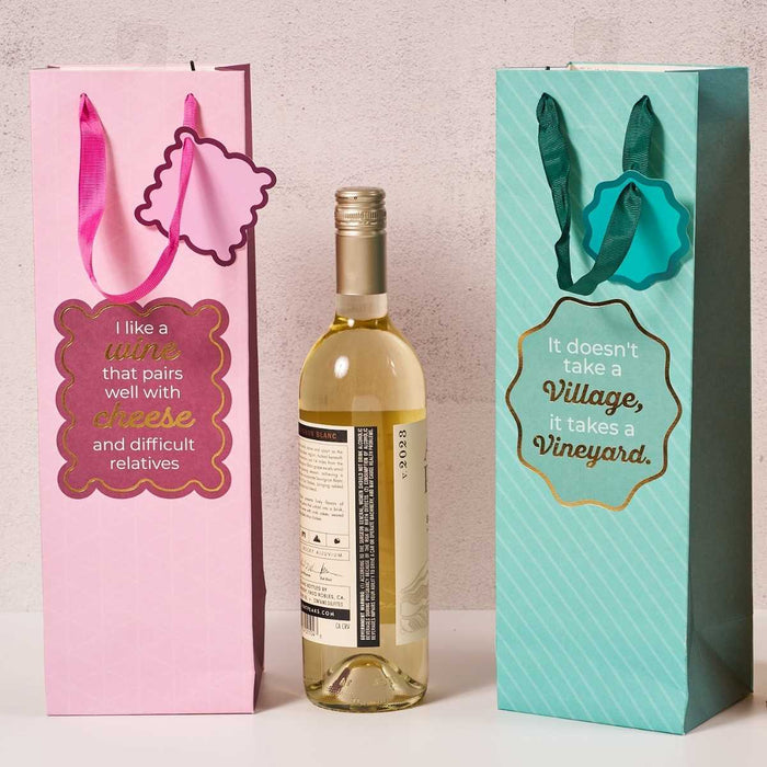 A bottle of white wine is flanked by two colorful Drinks On Me Vineyard Wine Bags—one pink with “I like a wine that pairs well with cheese and difficult relatives,” and one teal with “It doesn't take a village, it takes a vineyard.” Perfect hostess gifts.