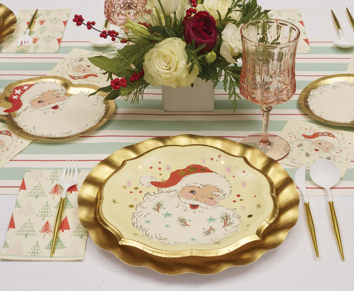 A festive table includes Sophistiplate Vintage Santa Dinner Plates (8pk), gold scalloped chargers, holiday napkins, a glass goblet, and a centerpiece of red and white flowers with greenery.