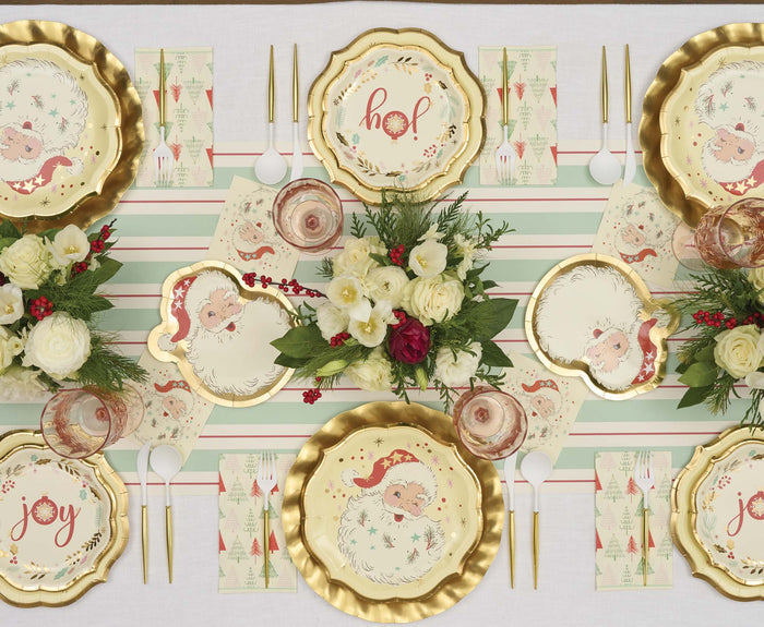 A festive Christmas table set with Sophistiplate Vintage Santa Dinner Plates (8pk) featuring joyful phrases, a striped pastel runner, white and red flowers, gold utensils, and holiday-themed napkins for a merry celebration.