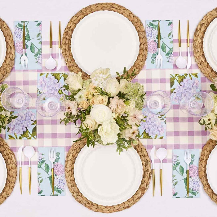 Create a spring-inspired Violet Hydrangea Table Setting by Sophistiplate with braided placemats, designer disposable plates, gold cutlery, floral napkins, and white and pale pink flowers atop a pink gingham runner.