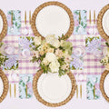 Create a spring-inspired Violet Hydrangea Table Setting by Sophistiplate with braided placemats, designer disposable plates, gold cutlery, floral napkins, and white and pale pink flowers atop a pink gingham runner.