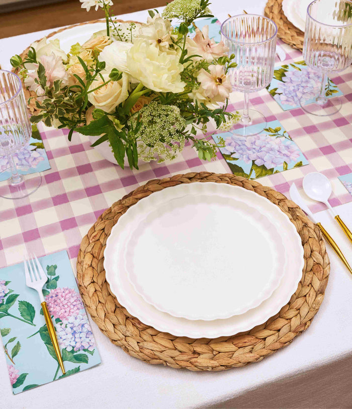 The Ultimate Easter Tablescape for 16: Violet Hydrangea by Sophistiplate LLC features woven placemats, designer plates, gold cutlery, floral napkins, and clear glasses on a pink gingham tablecloth with a pastel hydrangea centerpiece.