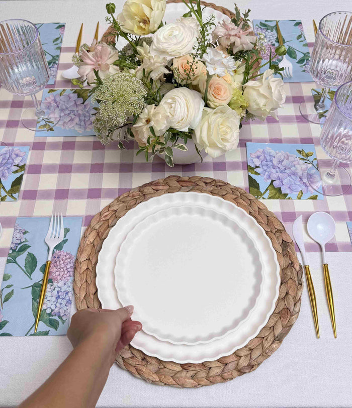 A hand sets a Sophistiplate Ultimate Easter Tablescape for 16: Violet Hydrangea designer plate on a table with floral napkins, gold utensils, clear glasses, and white and pale pink flowers atop a lavender checkered runner.
