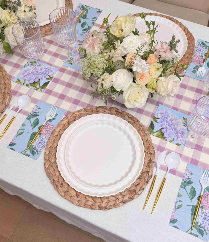 The Ultimate Easter Tablescape for 16: Violet Hydrangea by Sophistiplate LLC features woven placemats, designer disposable plates, gold and white cutlery, floral napkins, clear glasses, and a white and pastel centerpiece on a pink checkered cloth.