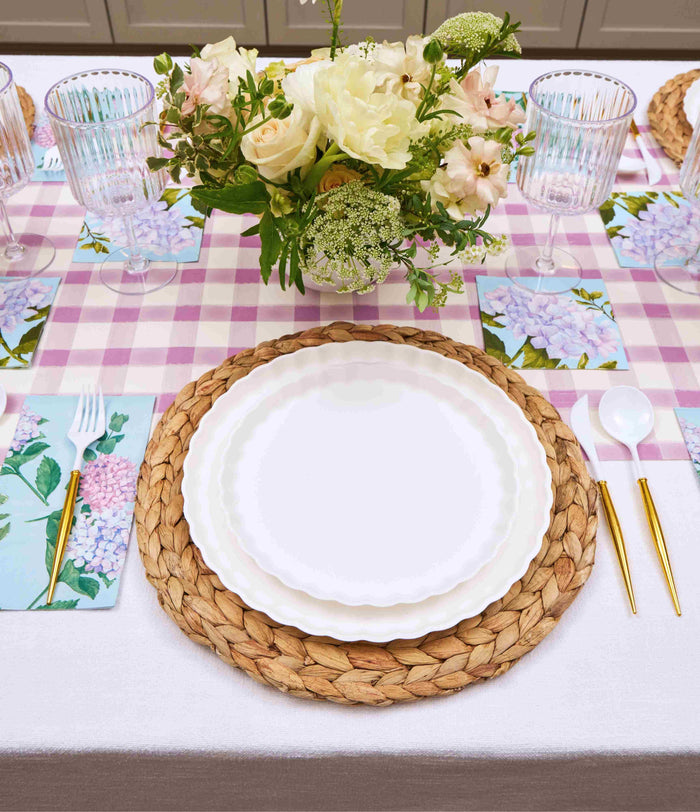 The Ultimate Easter Tablescape for 16: Violet Hydrangea by Sophistiplate LLC features woven placemats, designer disposable plates, gold flatware, floral napkins, ribbed glasses, and a white and pale pink floral centerpiece on a pink check runner.