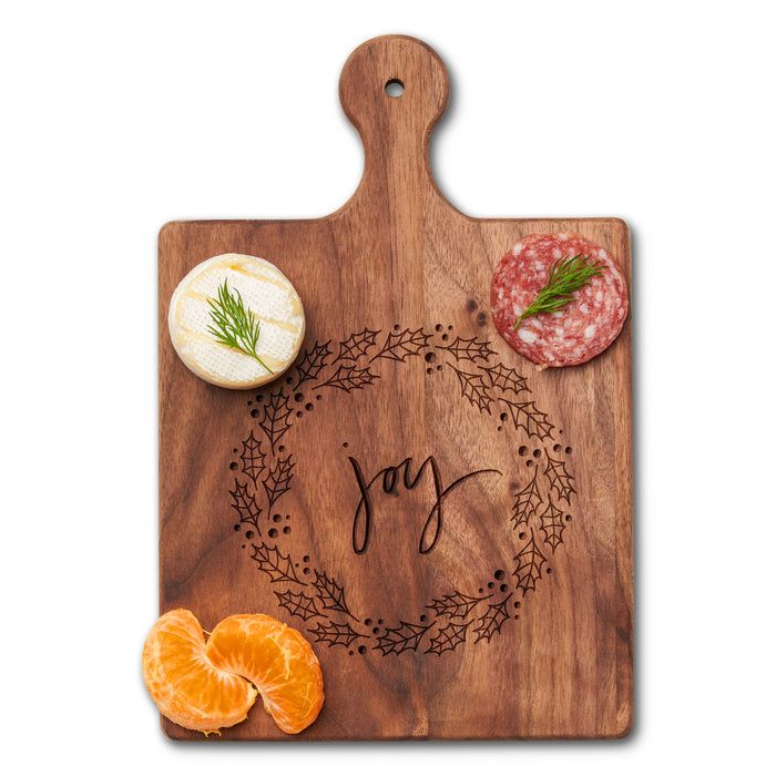 The Maple Walnut Artisan Paddle Board | Joy Wreath (9" x 6") showcases a joyful wreath engraving. It displays a round of cheese, dill salami, and orange segments neatly arranged on the elegant black walnut surface.