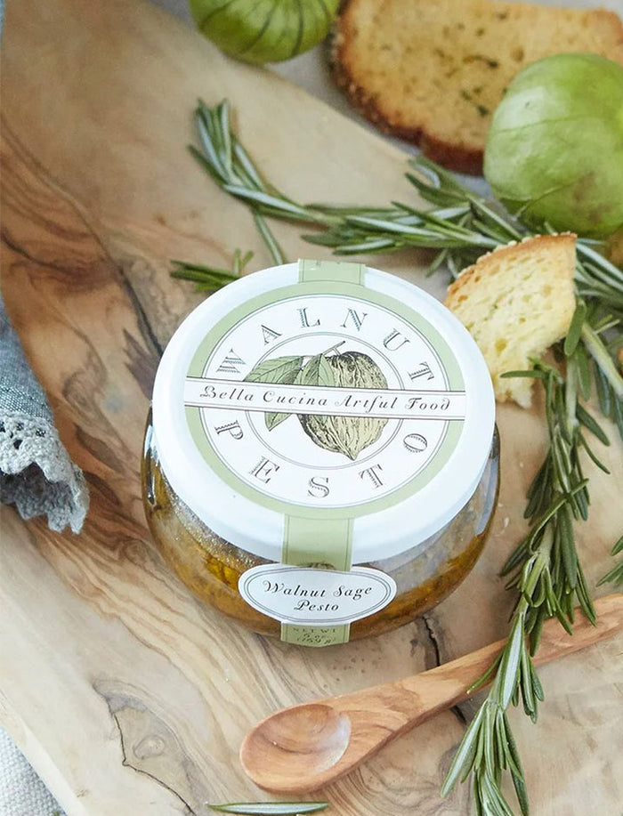 A jar of Bella Cucina Walnut Sage Pesto sits on a wooden board with fresh rosemary, Parmesan cheese, toasted bread slices, and green tomatillos. The decorative lid and packaging display "Walnut Sage Pesto" by Bella Cucina.