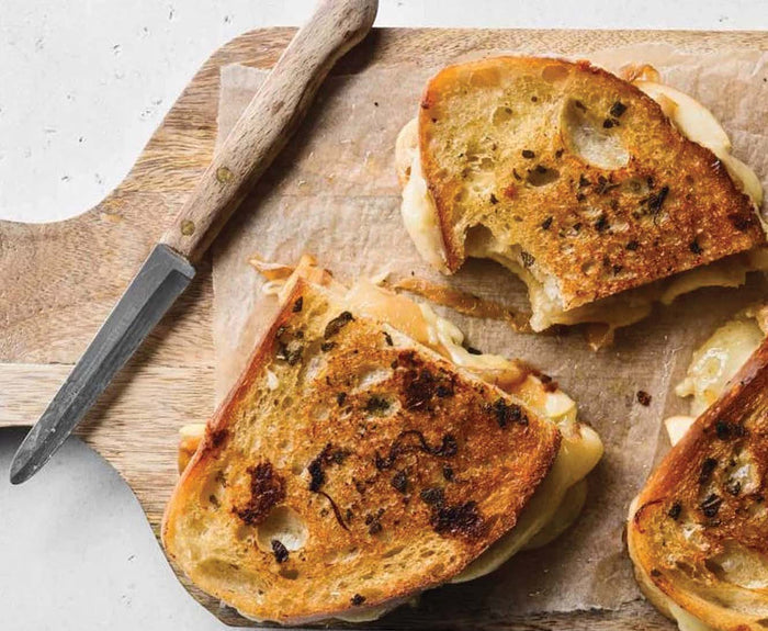 A close-up of grilled cheese sandwiches on a wooden board, one with a bite taken out, beside a small wooden-handled knife, featuring melted Parmesan cheese and Bella Cucina Walnut Sage Pesto.