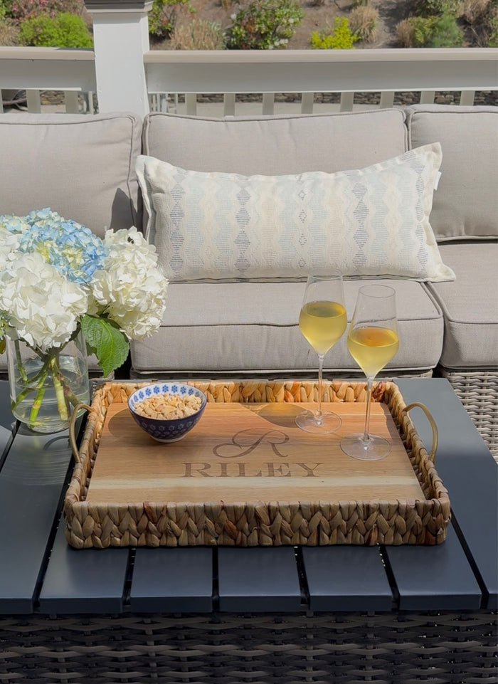 A Maple Water Hyacinth Rectangle Tray with Metal Handles and an Acacia Wood Liner, personalized with "RILEY," holds two white wine glasses and a small bowl on a patio table. White hydrangeas and a beige sofa with patterned pillows sit behind.