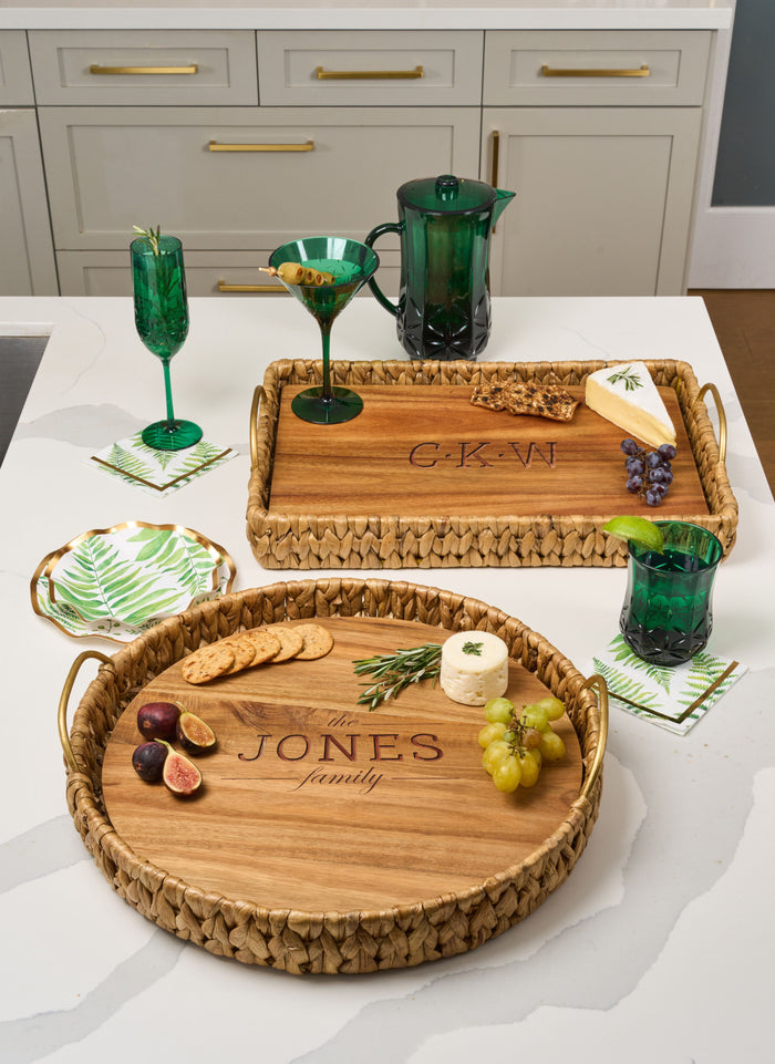Two Maple Water Hyacinth Rectangle Trays with metal handles and acacia wood liners, personalized, hold cheese, grapes, figs, crackers, and green glassware on a white counter.