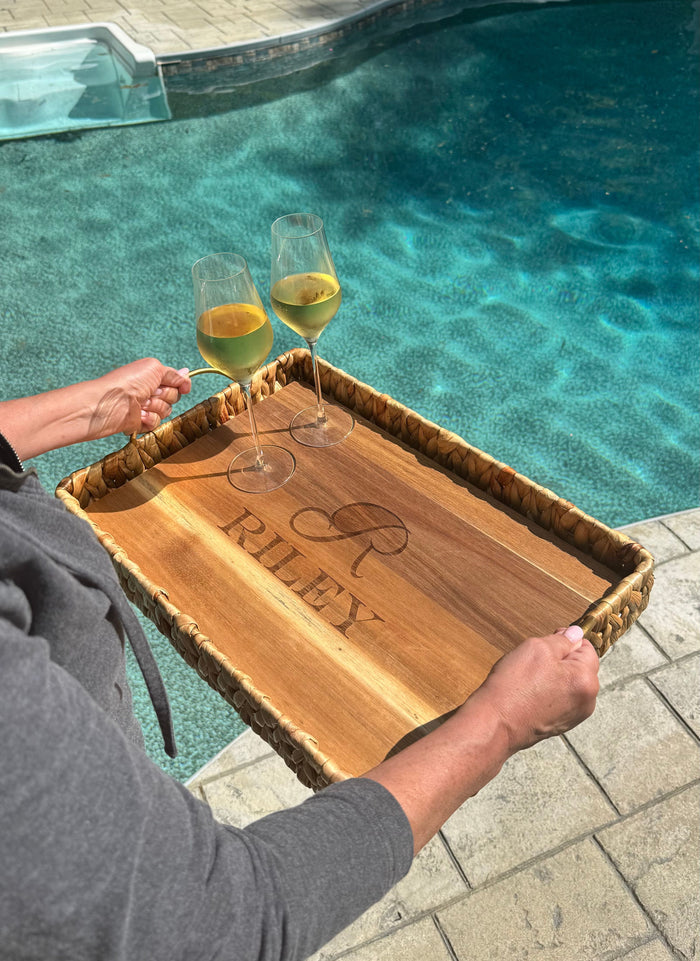 By the pool in bright sunlight, someone carries two glasses of white wine on a personalized Maple Water Hyacinth Rectangle Tray with metal handles and an acacia wood liner, measuring 19.5 x 13.5 x 4.5 inches.