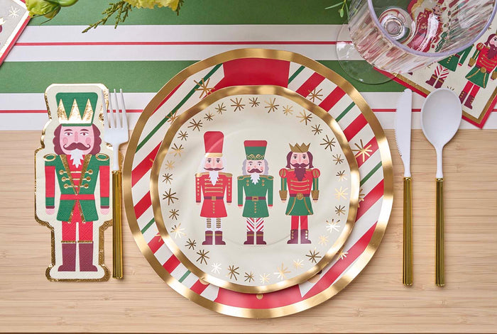A festive table setting features Sophistiplate's Wavy Salad Plate Nutcracker Christmas (8/pkg), gold-accented cutlery, a nutcracker-shaped napkin, and red, green, and white striped placemats.