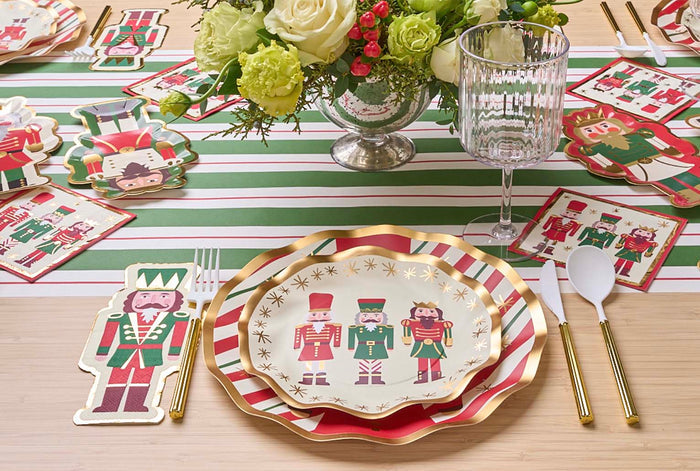 Create a festive holiday tablescape with the Sophistiplate Nutcracker Christmas Table Setting, featuring nutcracker plates, napkins, placemats, gold cutlery, glassware, a floral centerpiece, and a red, green & white striped table runner.
