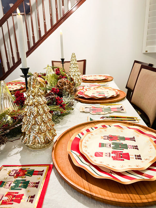A festive holiday table is set with Sophistiplate’s Wavy Salad Plate Nutcracker Christmas (8/pkg), gold and silver Christmas tree decorations, candles, and greenery on a white tablecloth by the staircase.