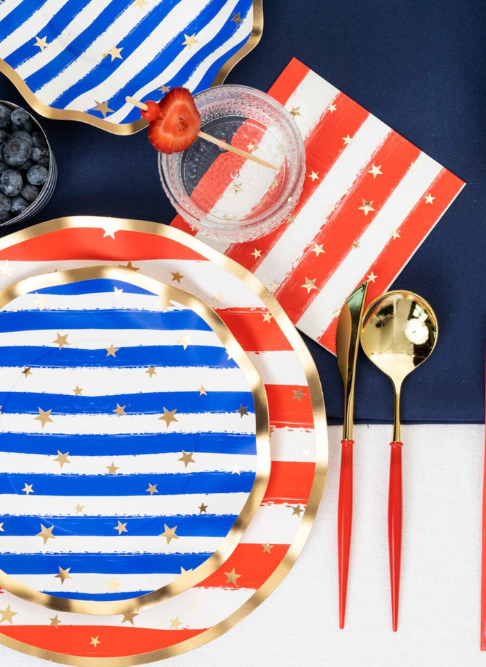 Create a festive table with Sophistiplate's Wavy Salad Plate Patriotic Confetti/8ct, gold utensils with red handles, a red and white napkin, a drink garnished with strawberry and blueberries—all set on a navy and white background.