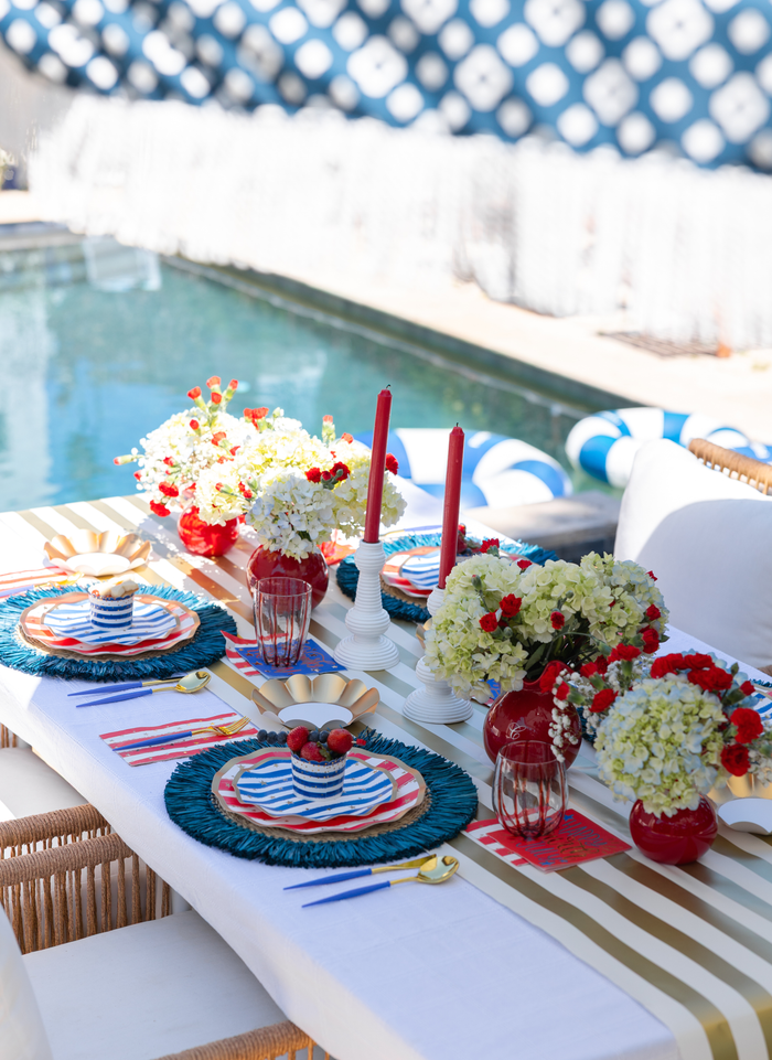 A festive outdoor poolside table is set with Sophistiplate LLC’s 4th of July Table for 16+: Patriotic Confetti, featuring colorful plates, blue placemats, gold-striped runners, red and white candles, and vibrant flowers in red and white vases.