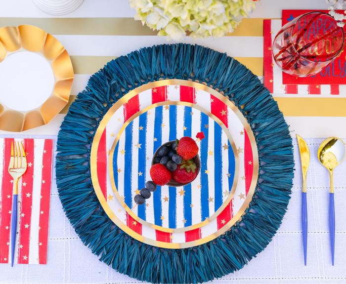Create a festive look with Sophistiplate LLC’s "4th of July Table for 16+: Patriotic Confetti" plates, adorned with gold stars and paired with gold utensils, berries, and red napkins on a white tablecloth.