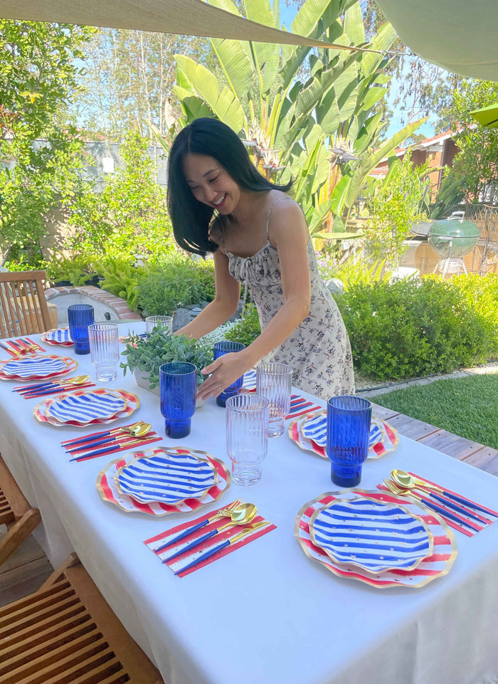 A woman in a floral dress sets a white outdoor dining table with Sophistiplate Wavy Dinner Plate Patriotic Confetti/8ct, gold utensils, and blue glasses, surrounded by lush greenery and wooden chairs.