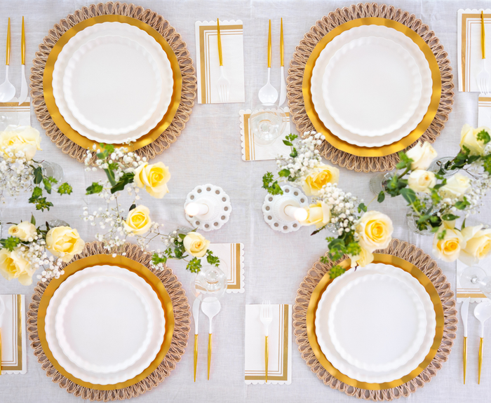 The Eco White & Gold Table Setting by Sophistiplate LLC features designer disposable plates, gold cutlery, and matching napkins on a white tablecloth, accented with yellow roses for a chic, symmetrical formal dining arrangement.
