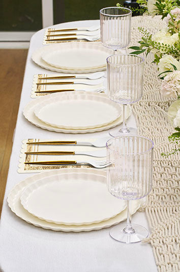 The Eco White & Gold Table Setting by Sophistiplate LLC features scalloped eco-friendly plates, gold flatware, ribbed clear glasses, and a light woven runner, all beautifully decorated with white and green floral arrangements.