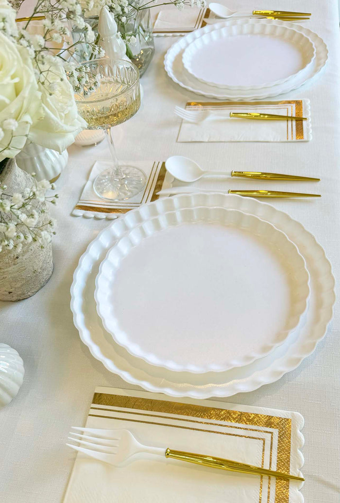 Create a chic tablescape with the Eco White & Gold Table Setting by Sophistiplate LLC, featuring scalloped eco-friendly plates, gold-handled cutlery, gold-trimmed napkins, floral centerpieces on a white tablecloth, and champagne glass.