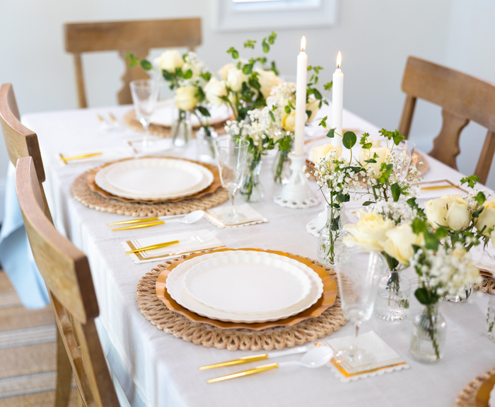The Eco White & Gold Table Setting by Sophistiplate LLC features eco-friendly plates, gold cutlery, woven placemats, lit white candles, and small vases of white flowers on a white tablecloth for an elegant dining experience.
