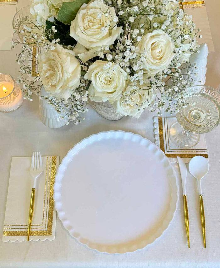 The Eco White & Gold Table Setting by Sophistiplate features biodegradable bamboo plates, gold-handled cutlery, gold-trimmed napkins, a glass, floral centerpiece of white roses and baby's breath on a white tablecloth, plus a lit candle.
