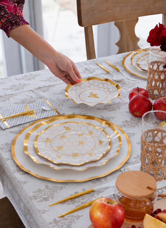 A hand sets a Sophistiplate White Honeybee Appetizer/Dessert Bowl on a table arranged with coordinating gold bee party plates, chopsticks, glasses, apples, and a floral-patterned tablecloth.