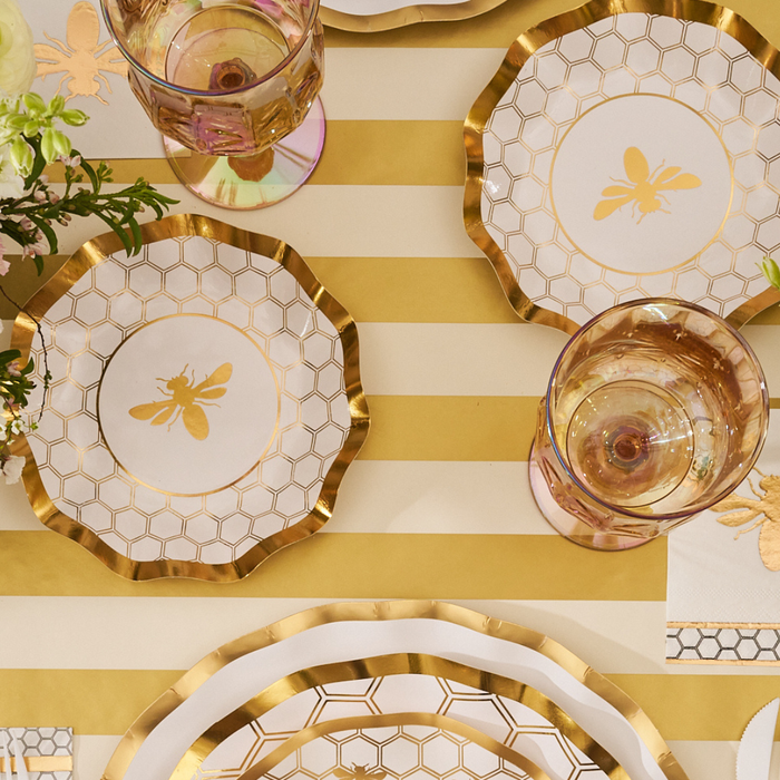 Gold and white table setting featuring Sophistiplate’s White Honeybee Appetizer/Dessert Bowls (8-pack), gold-rimmed glasses, and a floral arrangement atop a gold-striped tablecloth.