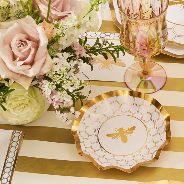 A set of Sophistiplate White Honeybee Appetizer/Dessert Bowls, featuring a charming gold bee and honeycomb design, is arranged on a gold-and-white striped table beside a pink and white flower bouquet and a pink-tinted glass—making these bowls perfect for adding a whimsical touch to your celebration.