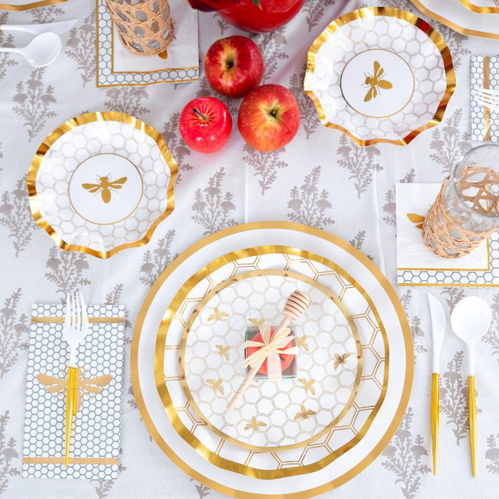 Table set with gold and white bee-themed plates, featuring Sophistiplate White Honeybee Appetizer/Dessert Bowls (8pk), gold bee party plates, apples, a honey dipper, and gold utensils on a patterned tablecloth. Decorative napkins, disposable paper bowls from Sophistiplate, and glasses complete the arrangement.