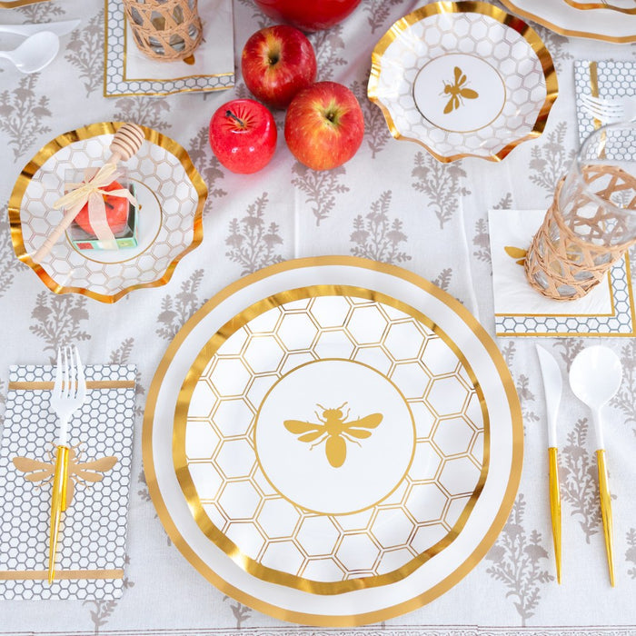A table set with Sophistiplate White Honeybee Dinner Plates (8pk), along with coordinating honeybee-themed napkins and cups, all accented in gold. Red apples and honey jars add vibrant color to the patterned white and gold plates atop a floral tablecloth.