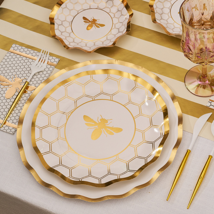 Sophistiplate’s White Honeybee Dinner Plates (8pk), featuring a charming honeycomb and bee motif with gold-rimmed, wavy edges, are arranged on a white and gold-striped tablecloth, complemented by gold flatware, a patterned napkin, and a pink-tinted wine glass for an effortlessly elegant tablescape.
