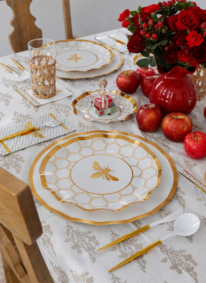 Create a stylish table arrangement featuring Sophistiplate White Honeybee Dinner Plates (8pk), gold cutlery, red apples, a vase of red roses, a glass with a woven sleeve, and a small candle-topped cake on a patterned tablecloth.