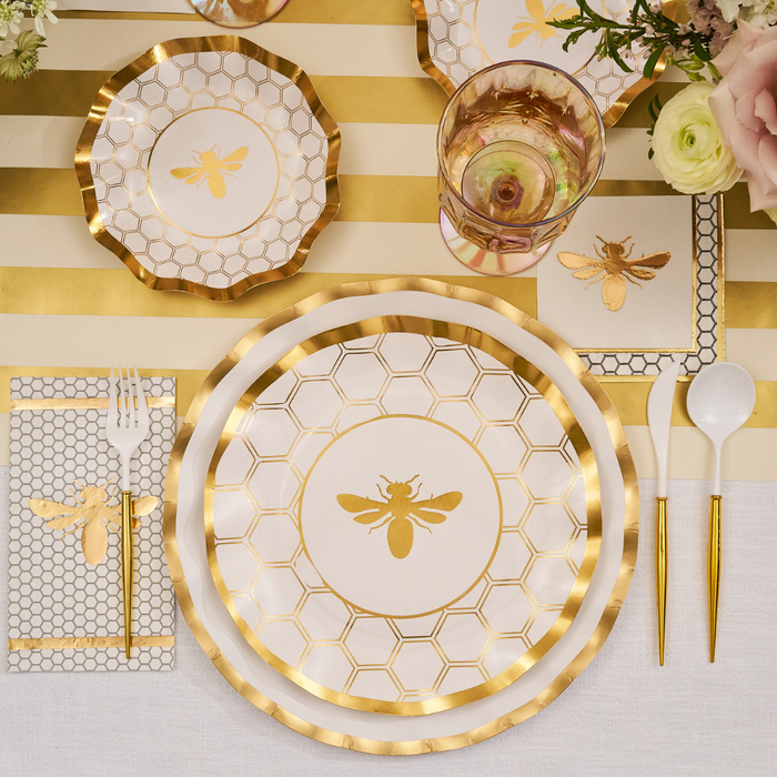 A table setting featuring Sophistiplate’s White Honeybee Appetizer/Dessert Bowls (8pk), gold and white plates with bee and honeycomb motifs, gold-rimmed utensils, a glass of pink beverage, and floral decorations atop a gold-striped tablecloth.