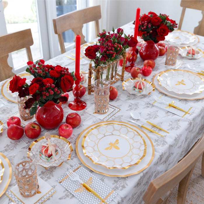 A dining table is set with Sophistiplate White Honeybee Dinner Plates, gold utensils, gold and white plates, red candles, red flowers in vases, pomegranates, and decorative napkins, creating an elegant and festive atmosphere.