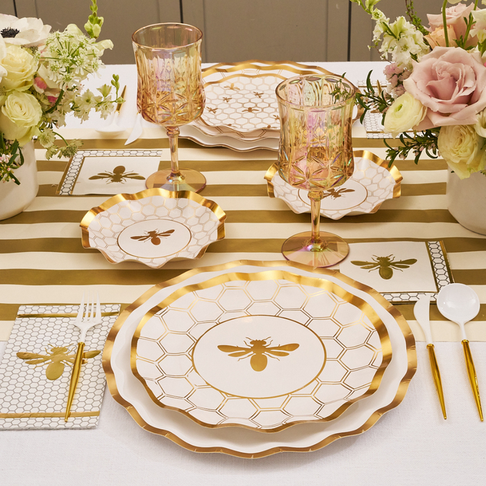 Create an elegant table setting featuring Sophistiplate’s White Honeybee Dinner Plates (8pk), wavy-edge dinner plates, a striped table runner, gold utensils, pink glasses, floral arrangements, and honeycomb-patterned napkins atop a crisp white tablecloth.