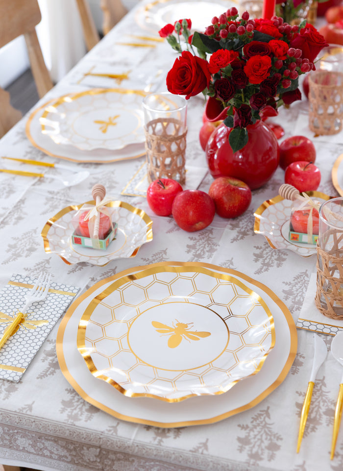 Elegant table setting featuring Sophistiplate White Honeybee Dinner Plates, honey dippers on small dishes, apples, red roses in a vase, and gold utensils atop a white patterned tablecloth.