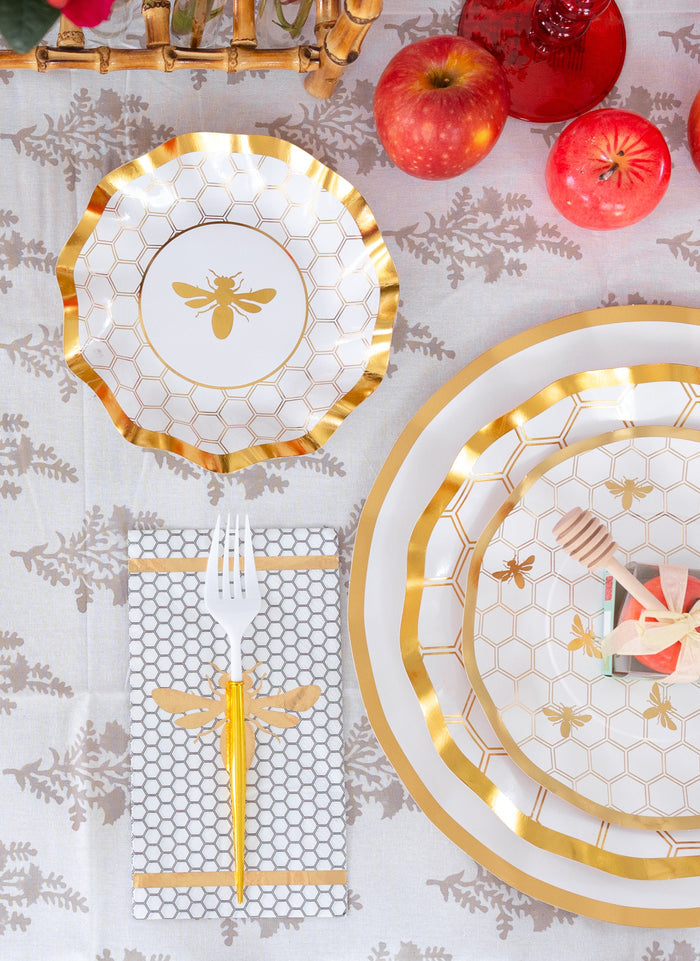 Sophistiplate’s White Honeybee Appetizer/Dessert Bowls (8pk) add elegance to the table setting, featuring intricate gold honeycomb and bee designs. The look is completed with a gold fork resting on a bee-patterned napkin, alongside apples and red drinks atop a white patterned tablecloth.