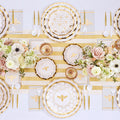 The Sophistiplate White Honeybee Table Setting features gold and white bee-themed plates, gold-rimmed paper plates, elegant gold utensils, a striped table runner, floral centerpieces with roses and ranunculus, and honeycomb-patterned napkins to create an elegant party tableware display.