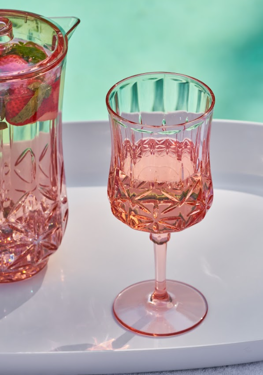 A Sophistiplate Wine Classic Stemmed | Blush | 16 Oz glass filled with a light pink drink sits on a white tray next to a matching pitcher with fruit and mint, set against a turquoise background evoking poolside vibes.