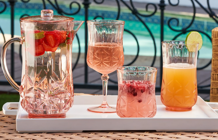 A glass pitcher of strawberry-infused drink and three Sophistiplate Tumbler Classic Tall | Blush | 21 Oz shatterproof tumblers, one with a lime slice, sit on a table by the pool, with decorative railing in the background.