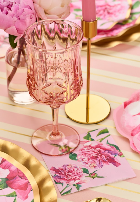The Sophistiplate Wine Classic Stemmed | Blush | 16 Oz sits on a striped pink and cream tablecloth beside a gold candle holder, floral napkin, and pink plates with gold trim. A small vase of pink flowers is nearby.