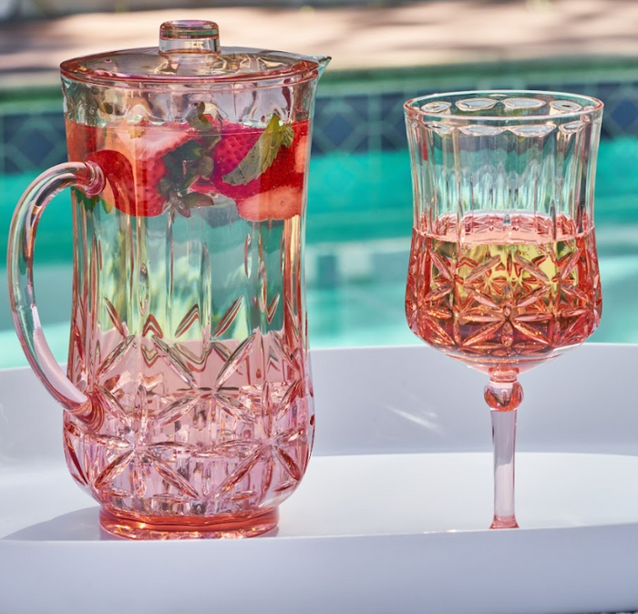 A pink glass pitcher with strawberry and mint-infused water sits next to a Sophistiplate Wine Classic Stemmed | Blush | 16 Oz BPA-free plastic wine cup on a white tray, with a blurred swimming pool in the background.