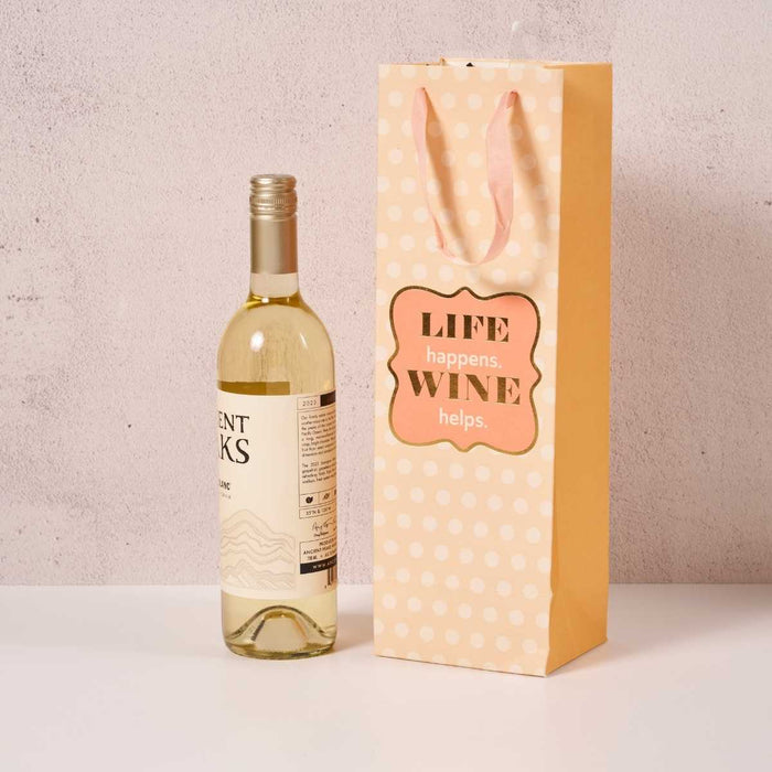 A bottle of white wine sits beside the Drinks On Me Wine Helps-Wine Bag, a yellow polka-dot gift bag with pink handles and “LIFE happens. WINE helps” label—perfect for hostess gifts or special occasions.