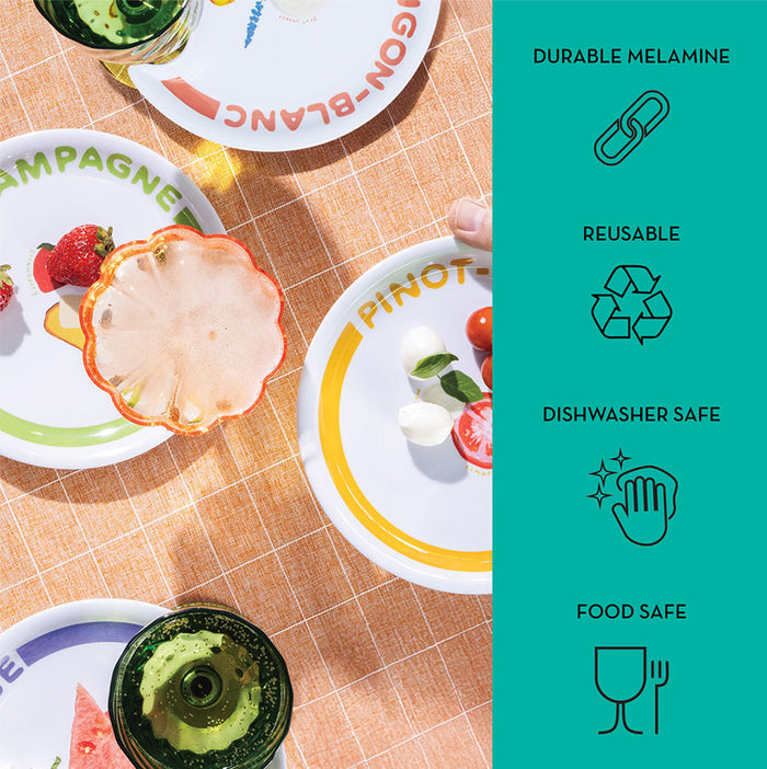Maple’s Wine Holder Appetizer Plates by Meg Quinn—colorful melamine plates with fruit and veggie designs—rest on a peach-checkered tablecloth. A teal panel notes they’re durable, reusable, and dishwasher safe with food-safe icons.