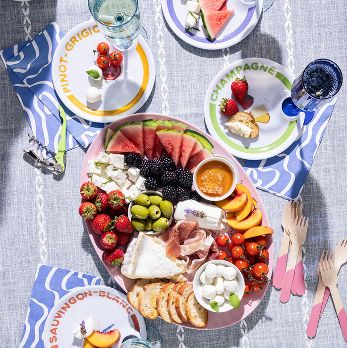 A vibrant picnic on a blue tablecloth featuring Maple’s Wine Holder Appetizer Plates by Meg Quinn, filled with charcuterie, cheeses, fruit, bread, prosciutto, olives, honey—and crafted slots for glasses of wine and water.