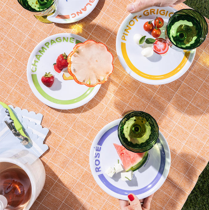 A table set for an outdoor meal showcases Maple’s Wine Holder Appetizer Plates by Meg Quinn, labeled CHAMPAGNE, PINOT GRIGIO, and ROSÉ, each with fruit and cheese. Hands reach for plates on a peach checkered tablecloth.